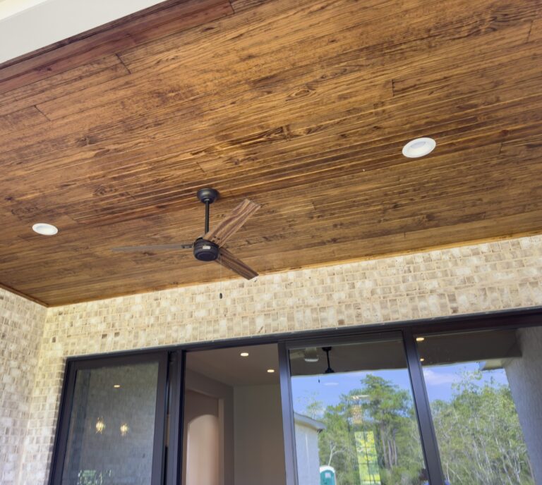 Covered rear porch with a stained pine tongue-and-groove ceiling, ceiling fan, recessed lighting, light brick exterior wall, and sliding glass doors at the Cypress custom home in Florence Gardens, Gulfport, Mississippi.