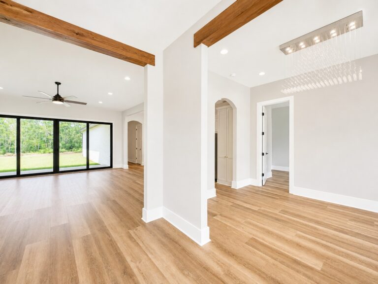 Open foyer and living area with cypress wrapped beams, wood-look flooring, modern chandelier, and large sliding glass doors in the Cypress custom home at 12483 Alphabet Road in Florence Gardens, Gulfport, Mississippi 39503.