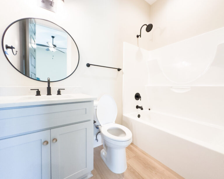 Downstairs guest bathroom with a blue-gray vanity, round mirror, tub-and-shower combination, matte black fixtures, and light wood-look flooring at 12487 Alphabet Road in Florence Gardens, Gulfport, Mississippi 39503.