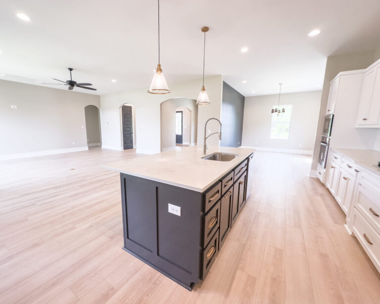 Open-concept kitchen, dining, and living area with a dark kitchen island, white cabinetry, pendant lighting, arched openings, and light wood flooring at 12487 Alphabet Road in Florence Gardens, Gulfport, Mississippi 39503.
