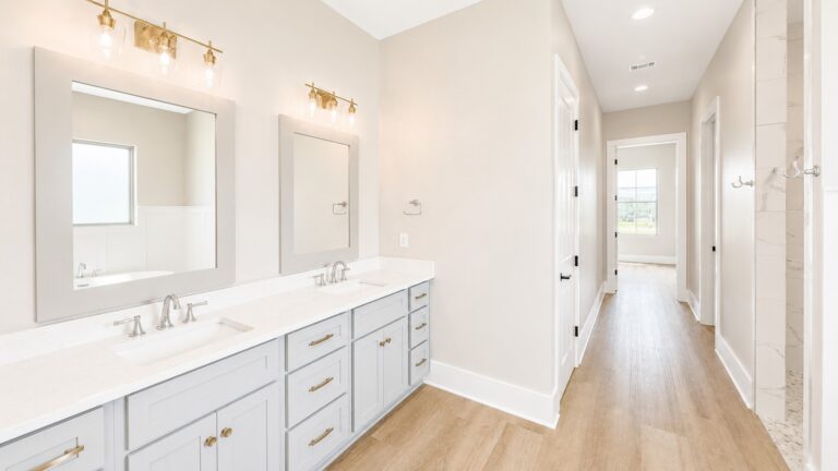 Primary bathroom with a long gray double vanity, white countertops, framed mirrors, brass lighting, and wood-look flooring in the Cypress custom home at 12483 Alphabet Road in Florence Gardens, Gulfport, Mississippi 39503.