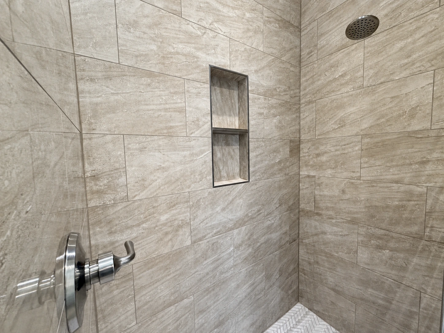 Walk-in shower with large beige tiles, recessed shelving niche, herringbone floor tile, and modern metal shower fixtures by BW Artisan Homes in Gulfport.