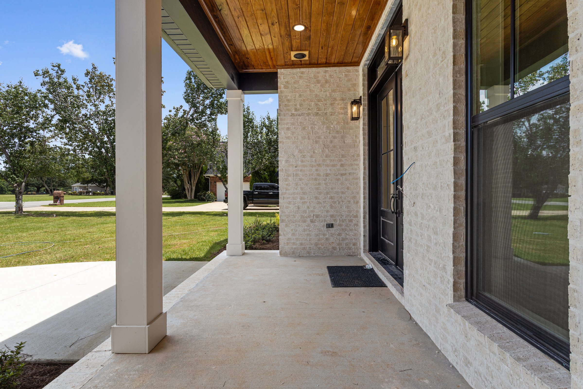 Covered porch with brick walls, black windows and door, wood ceiling, and wall lanterns by BW Artisan Homes in Gulfport, MS.