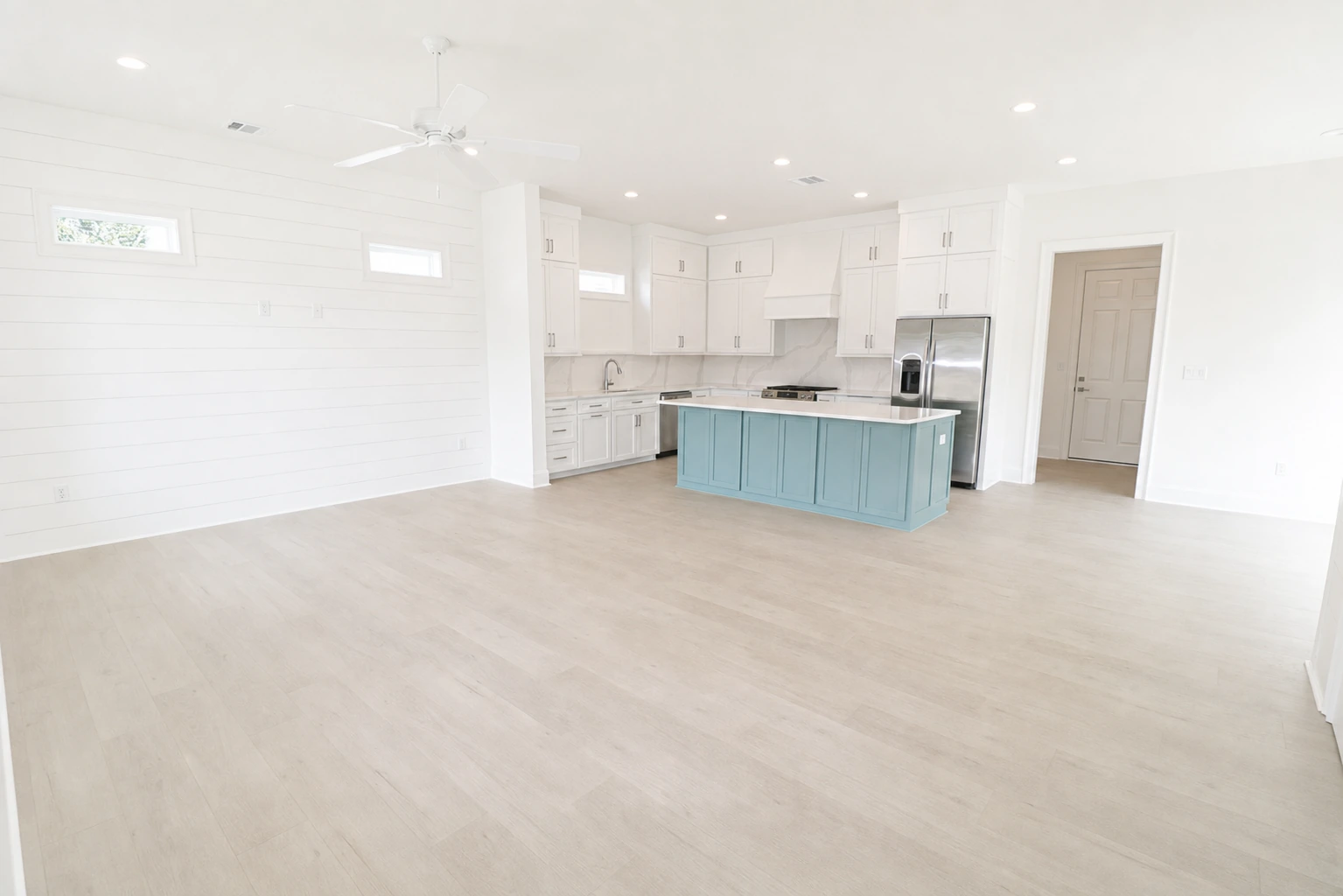 Bright open kitchen and living area with white custom cabinetry, blue kitchen island with quartz countertop, ceiling fan, and light wood floors by BW Artisan Homes in Ocean Springs