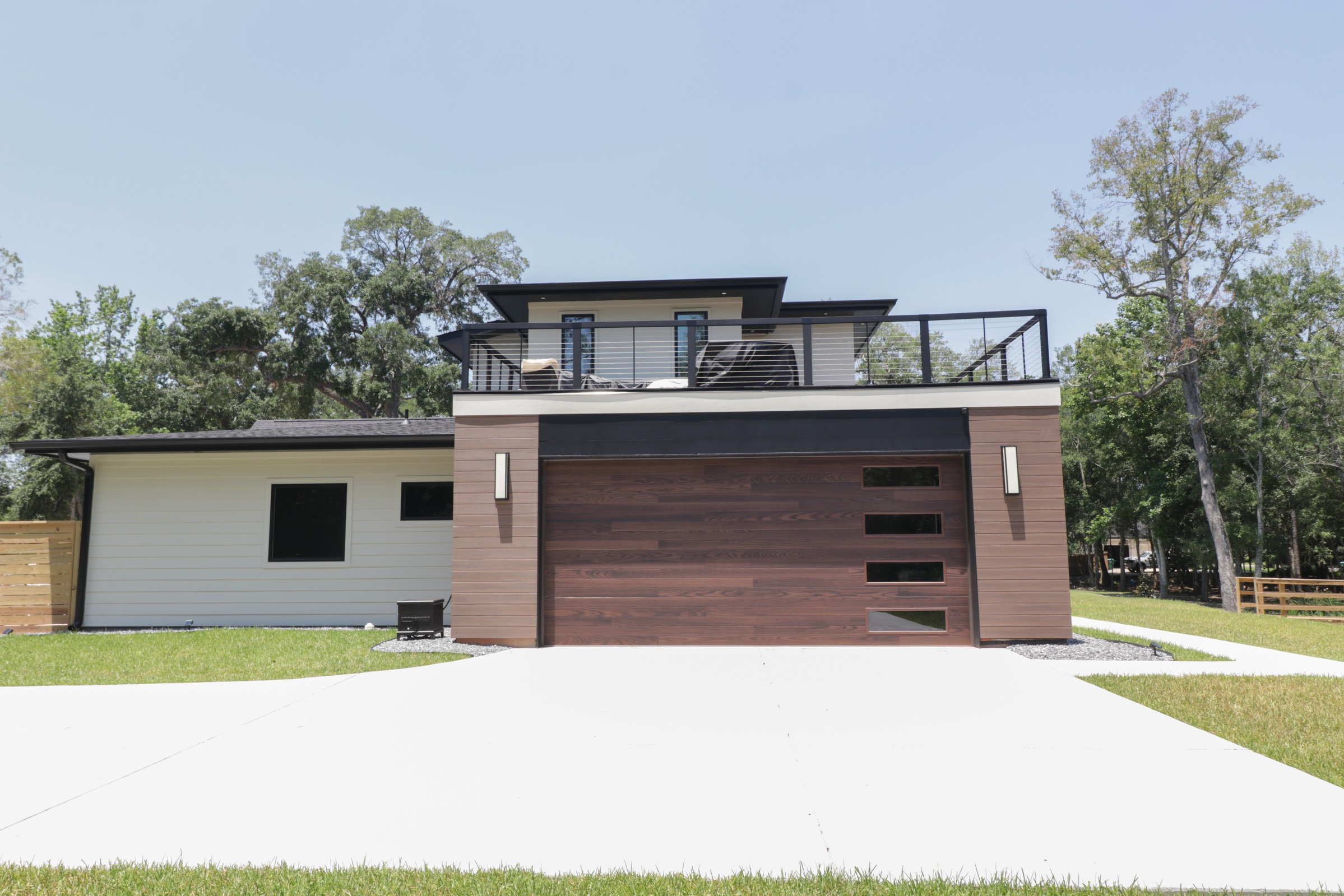 Modern home exterior featuring a wood-finish garage door, black windows, and rooftop deck with metal railing by BW Artisan Homes in Gulfport, MS.