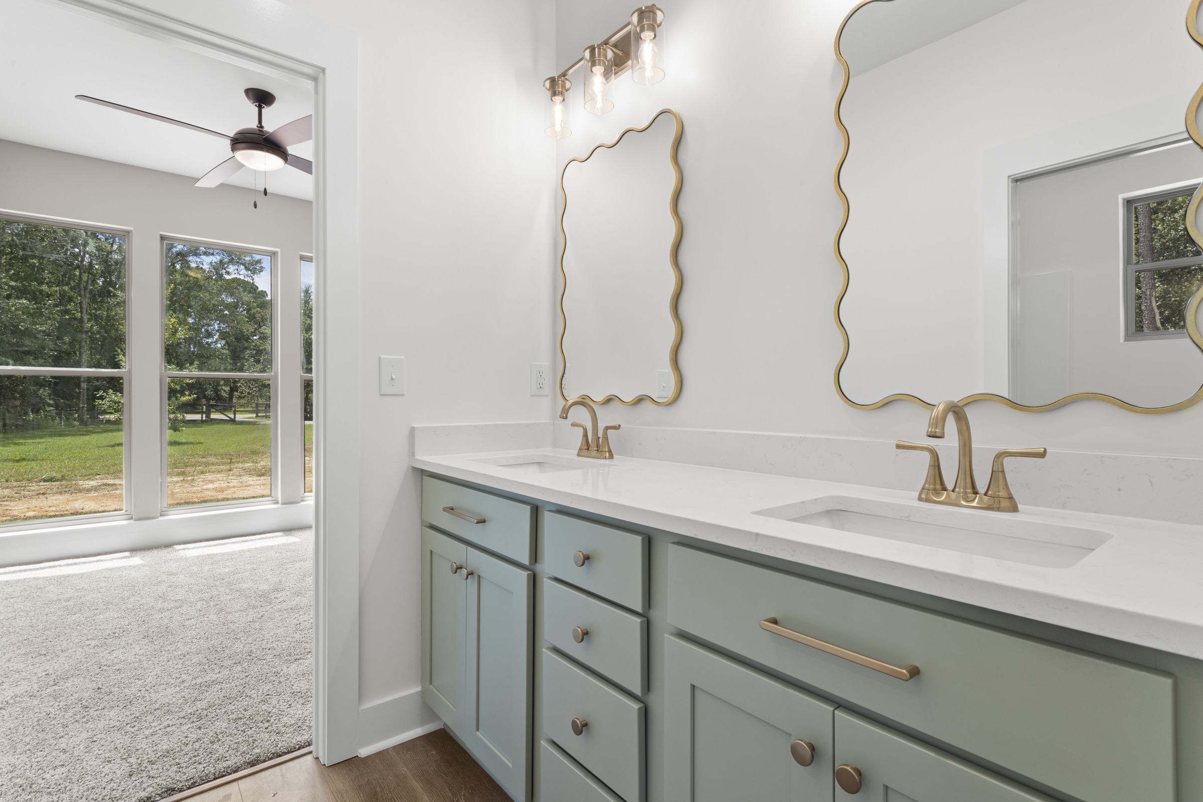 Bathroom vanity with dual sinks, quartz countertops, soft green custom cabinetry, decorative brass-framed mirrors, and large windows showing greenery outside, by BW Artisan Homes in Pass Christian.