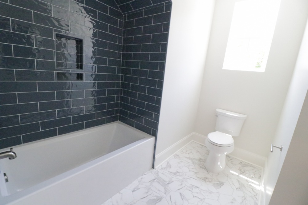 Bathroom with white bathtub, dark glossy subway tile wall, marble-look tile floor, toilet, and natural light from a high window in BW Artisan Homes, Ocean Springs
