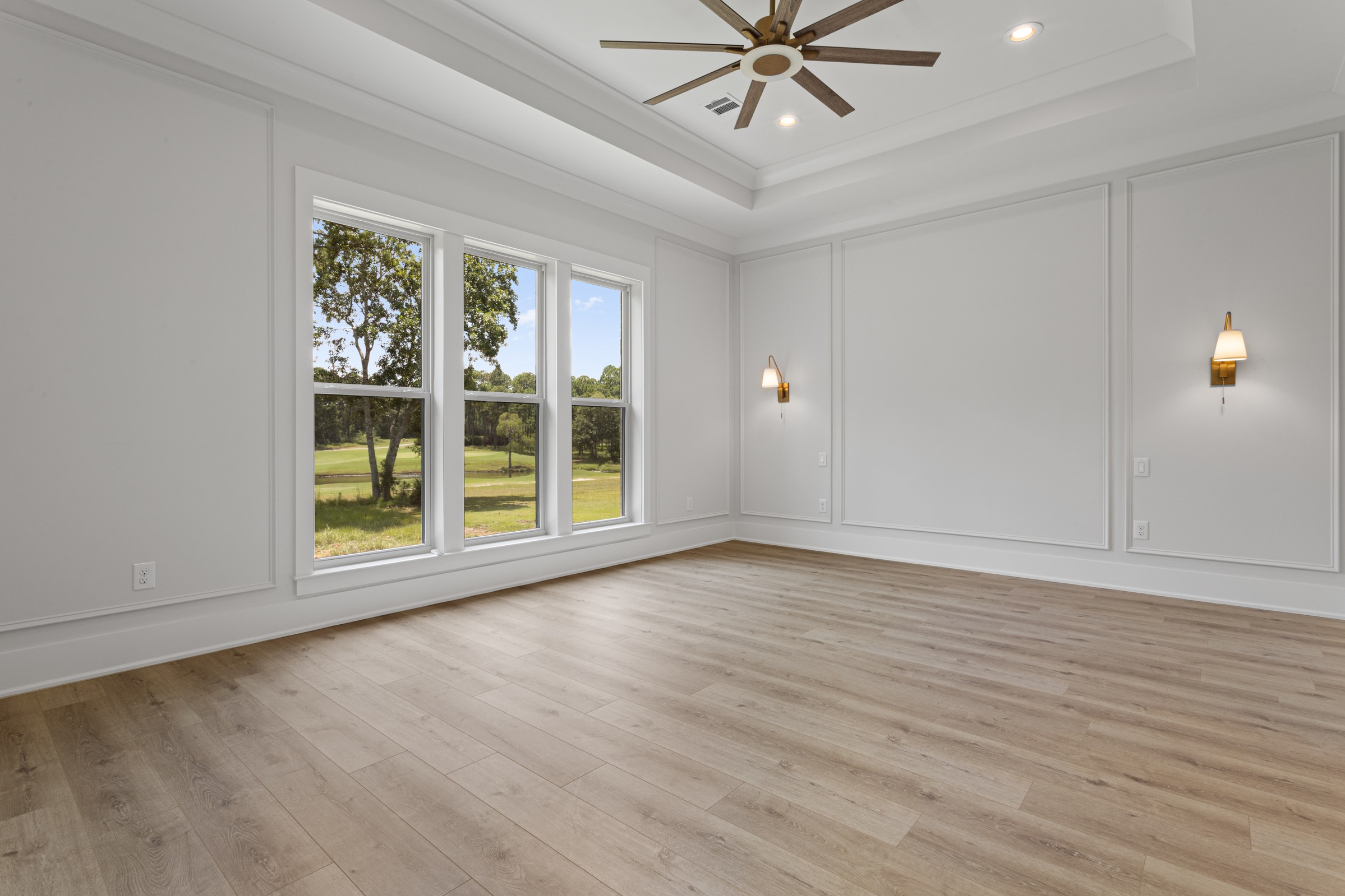 Bright bedroom with expansive windows overlooking greenery, white oak floors, tray ceiling with fan, and modern wall sconces by BW Artisan Homes in Gulfport, MS.