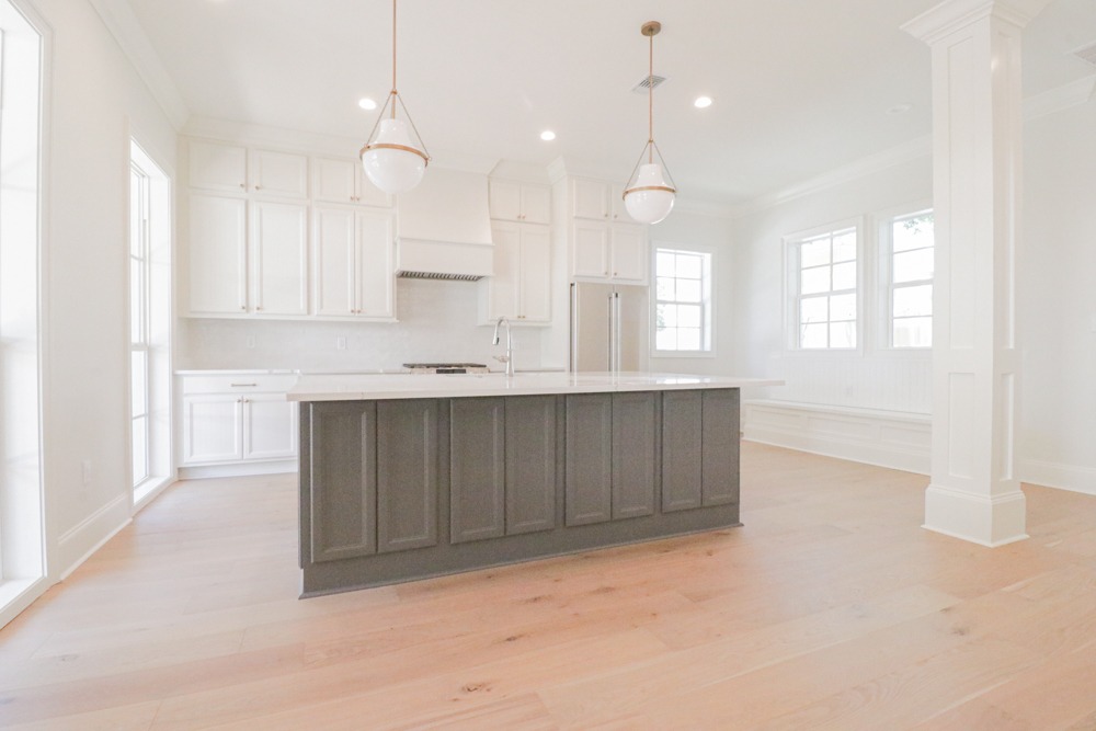 Bright kitchen with white custom cabinetry, dark kitchen island with quartz countertop, pendant lights, and white oak floors by BW Artisan Homes in Ocean Springs, MS.
