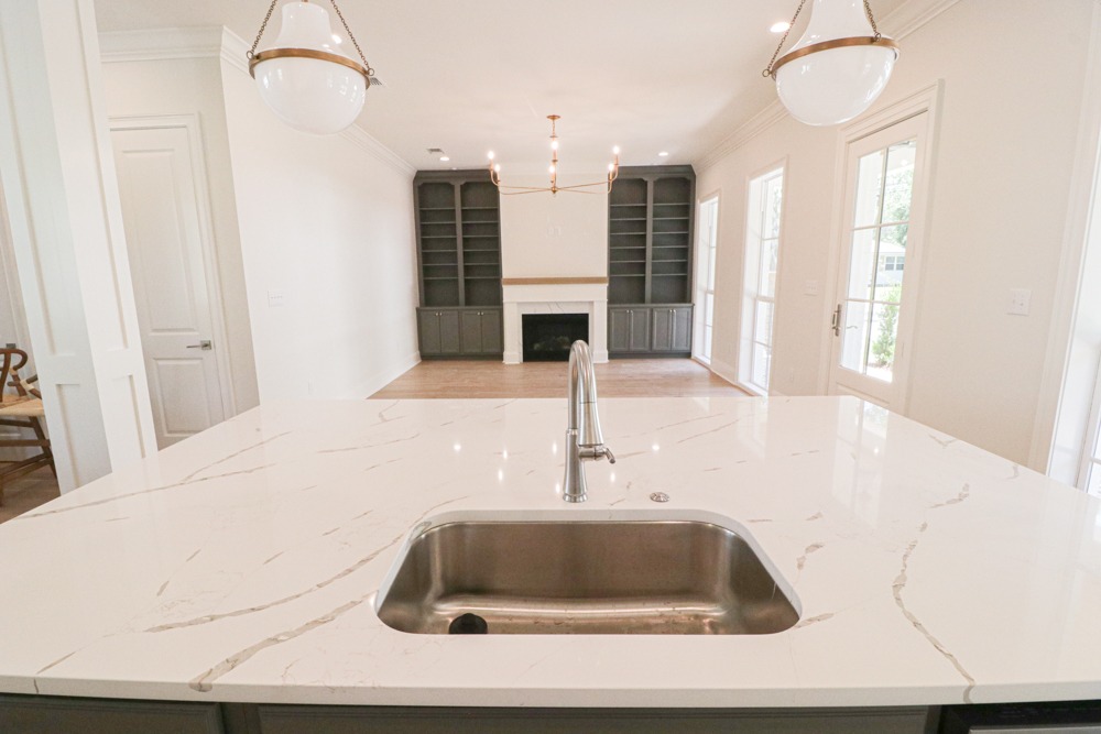Bright kitchen island with quartz countertop and stainless steel sink looking towards living room with built-in cabinetry and fireplace in Ocean Springs by BW Artisan Homes.